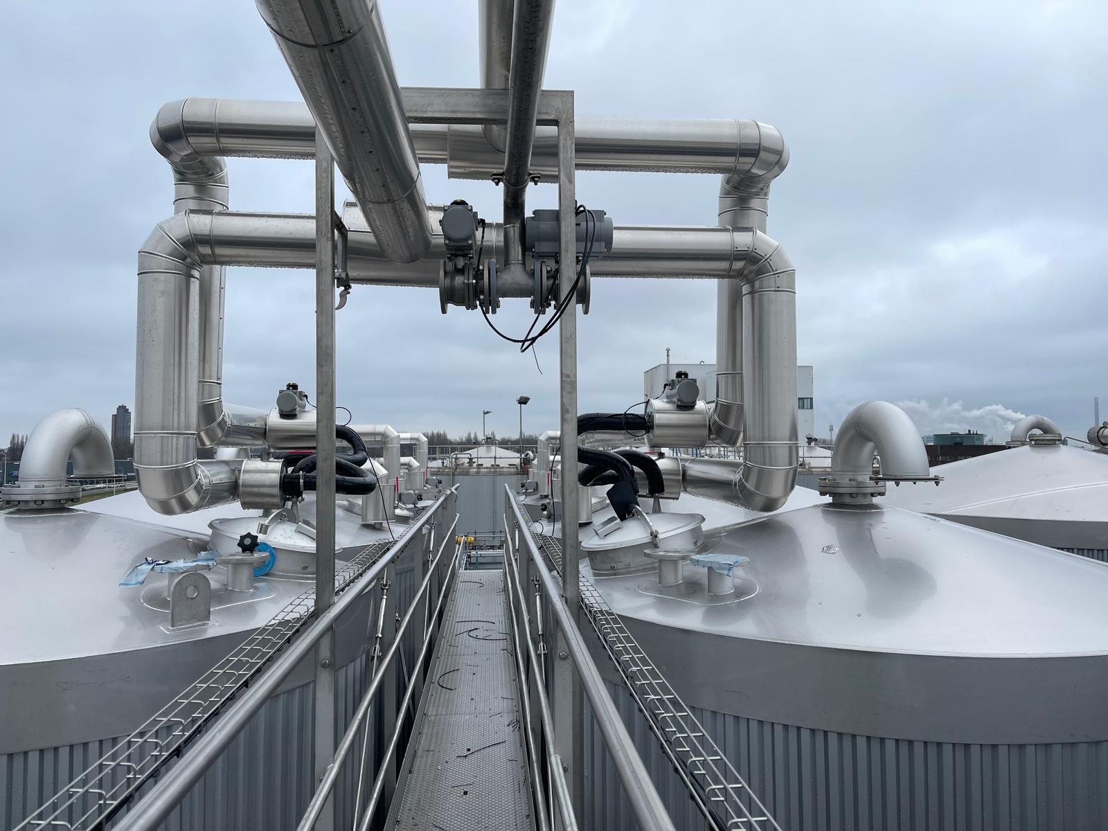Met aluminium beplaat procesleidingwerk op een stalen frame boven een rij geïsoleerde tankdaken, met een looppad ertussen