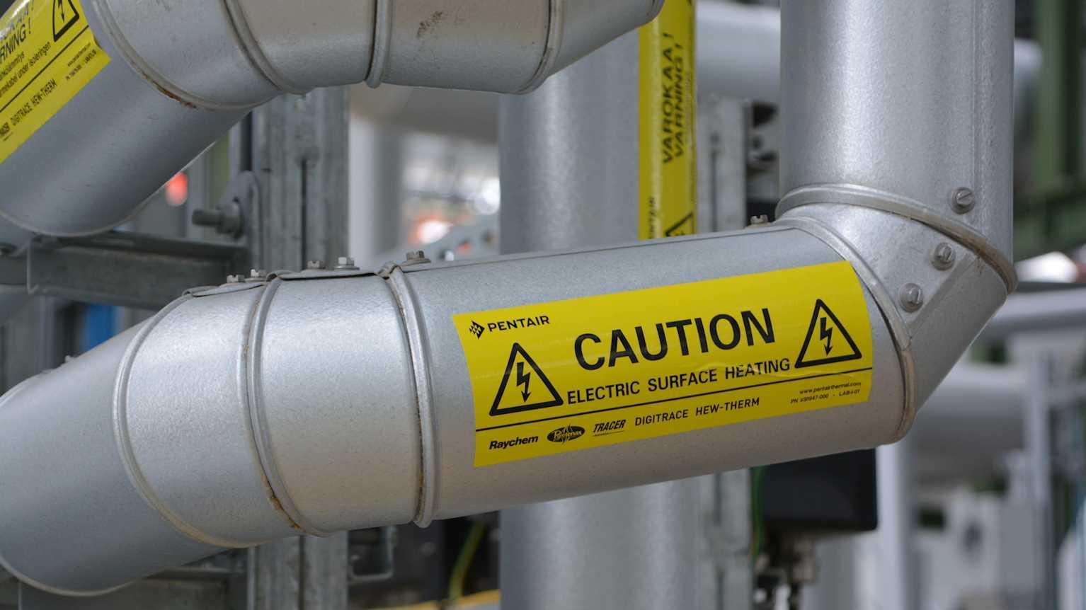Met aluminium beplaat leidingwerk met een geel waarschuwingslabel 'Caution – Electric surface heating' op de beplating