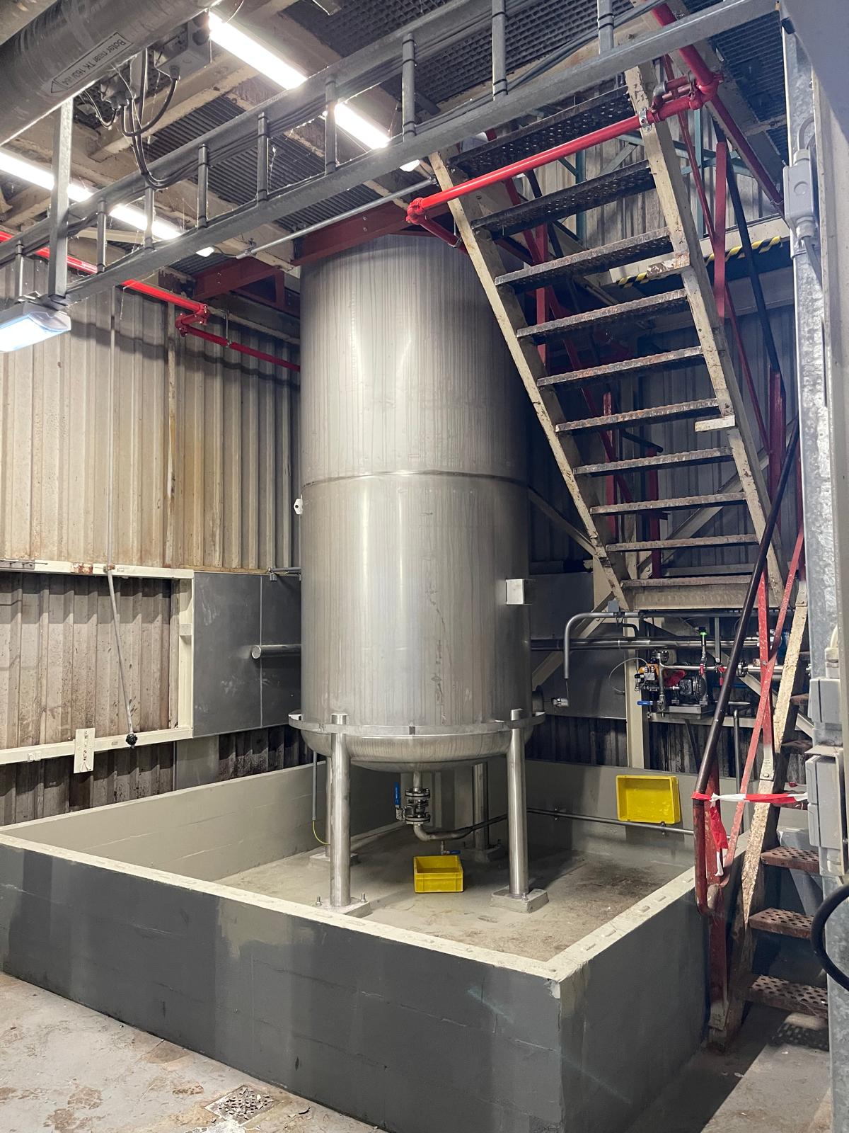 Ongeïsoleerde verticale tank op poten in een betonnen opvangbak, naast een stalen trap
