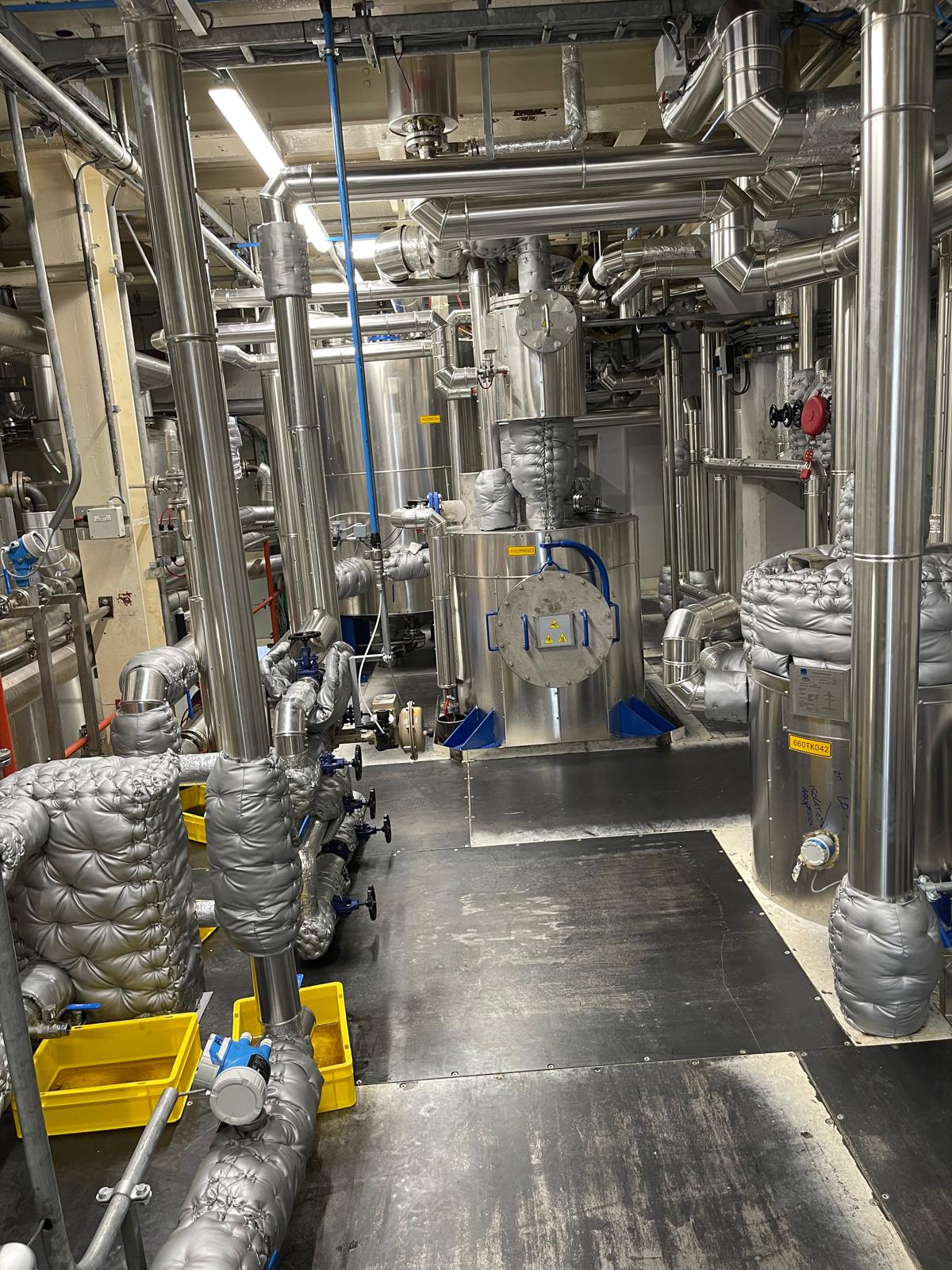 Technische ruimte met leidingwerk met RVS-beplating, verticale stijgleidingen en procesapparatuur, met grijze afneembare isolatiemantels op afsluiters en leidingdelen