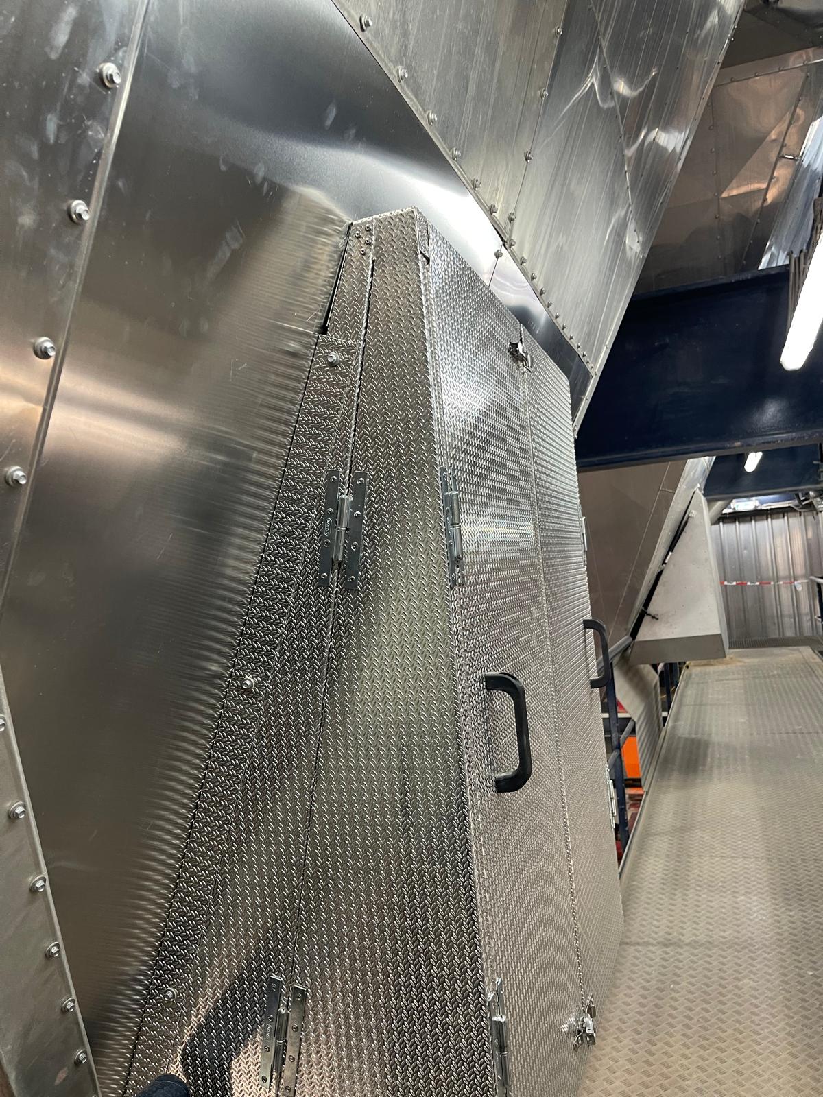 Zijaanzicht van een taps toelopende omkasting van aluminium tranenplaat met scharnieren en een deurgreep, bevestigd tegen geklonken aluminium beplating naast een looppad van tranenplaat