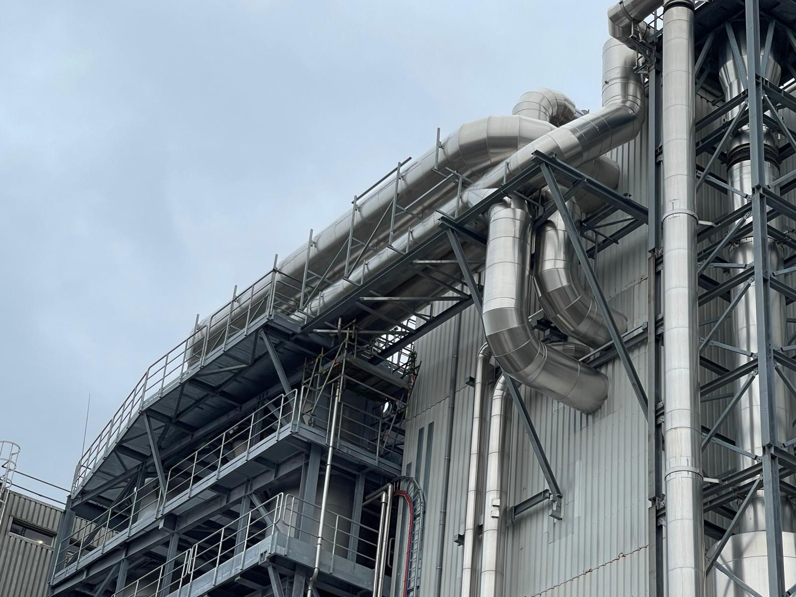 Grote geïsoleerde leidingtracés met afgewerkte metalen buitenbeplating op een staalconstructie naast een industrieel gebouw