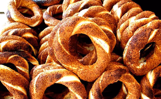 Pile of golden brown simit bread rings coated with sesame seeds on a black background.