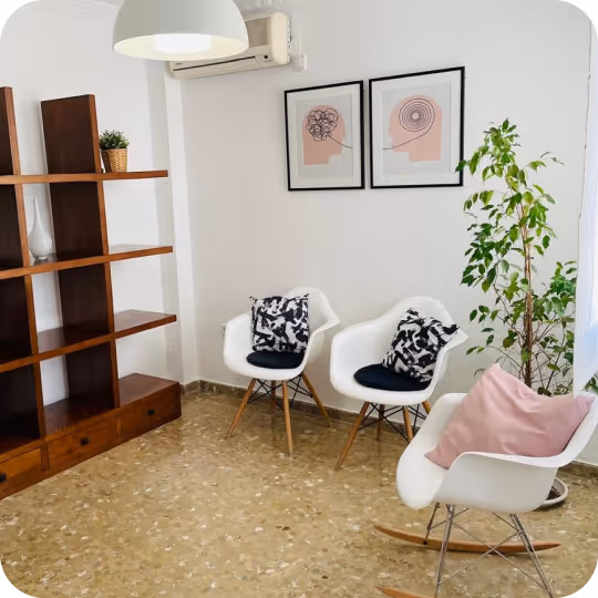 Otra imagen del consultorio del psicólogo: habitación moderna con dos sillas blancas con cojines negros, una mecedora blanca con un cojín rosa, una estantería de madera, dos obras de arte abstractas enmarcadas y una planta alta de color verde.