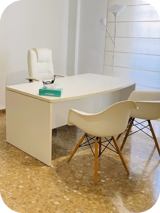 Imagen del consultorio del psicólogo: oficina minimalista blanca con un escritorio curvo, silla giratoria blanca, dos sillas para visitantes blancas con patas de madera y una lámpara de pie alta.