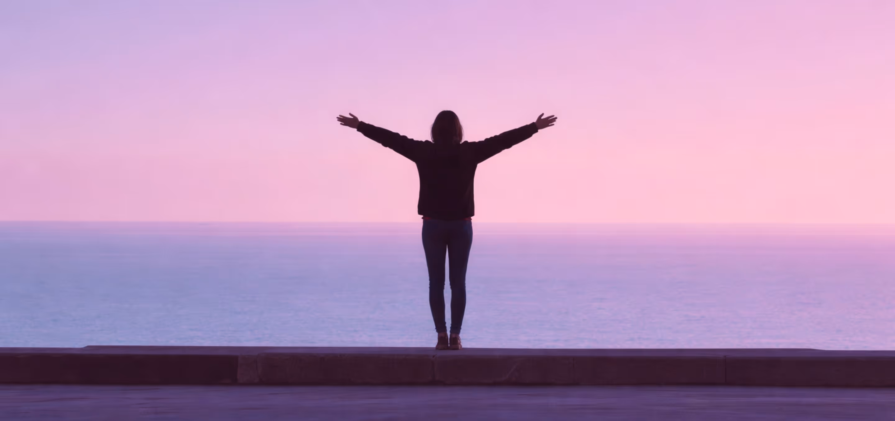 Silueta de una persona de pie con los brazos extendidos, mirando al océano bajo un cielo al atardecer de tonos rosa y púrpura.