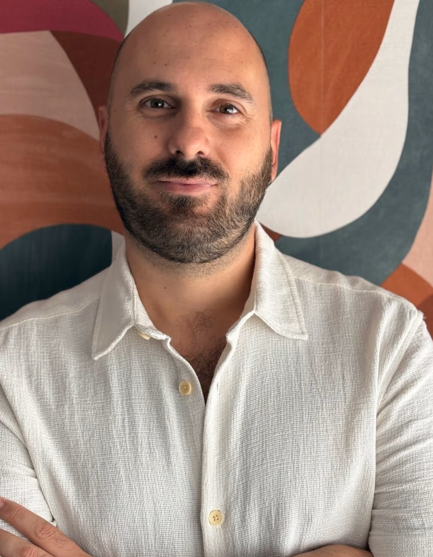 Primer plano del psicólogo, Rafael. Es calvo, con barba, viste una camisa blanca de botones con textura y está de pie con los brazos cruzados frente a una pared abstracta y colorida.