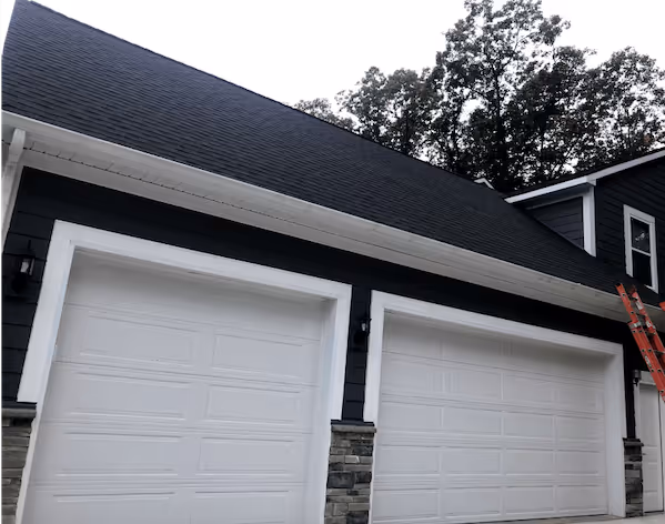 Double garage with white doors and black siding under a dark shingled roof with a red ladder leaning against the house.