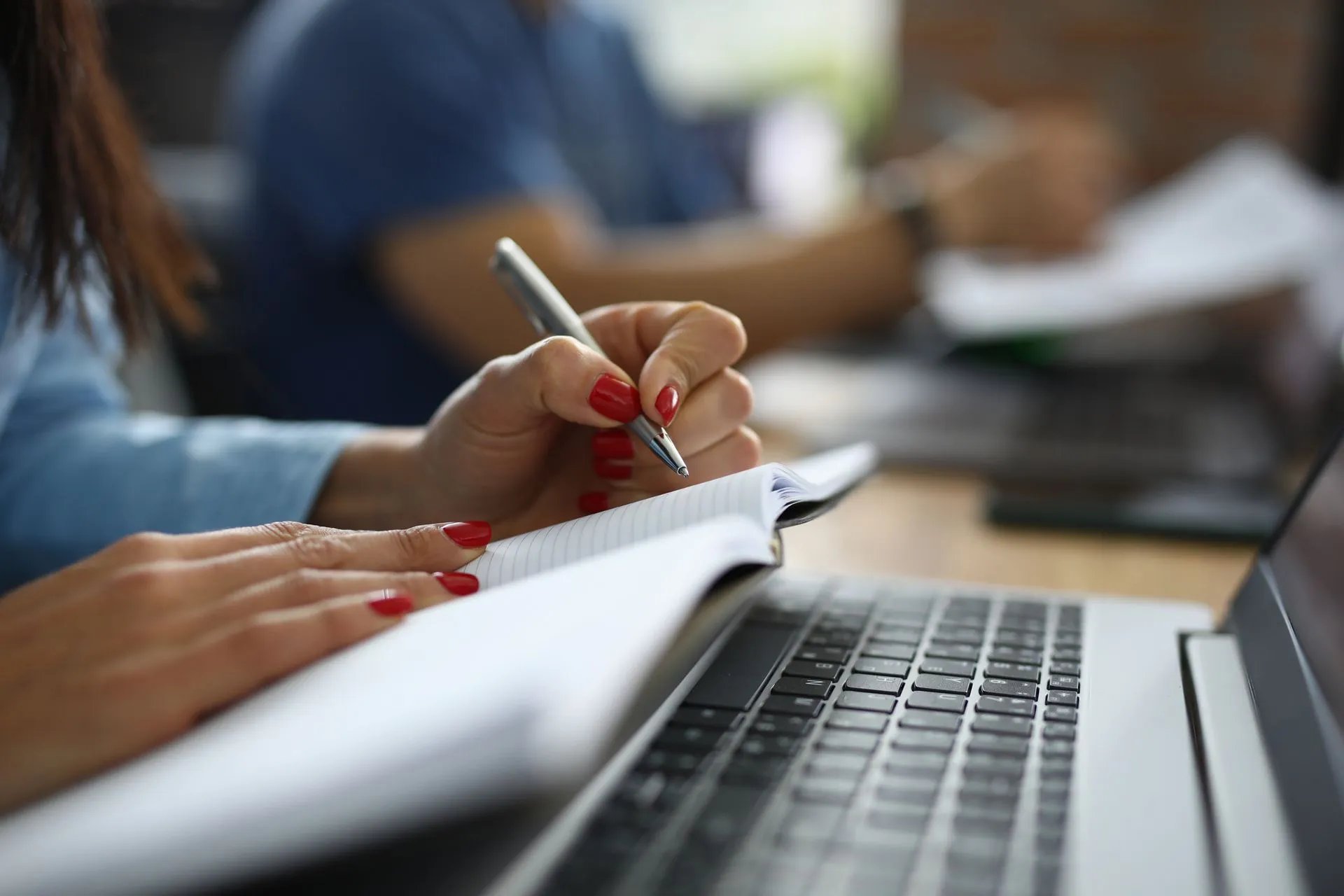Person with red nail polish writing in a notebook placed on a laptop keyboard.