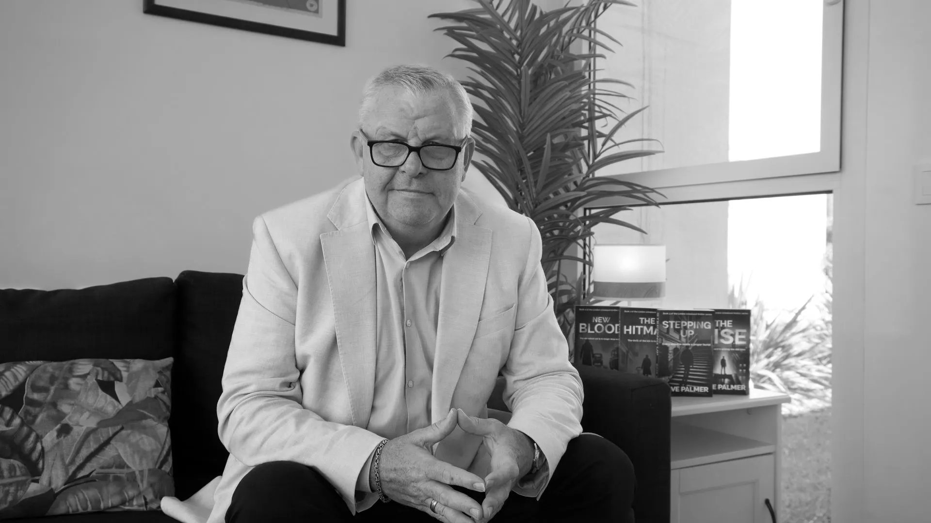 Older man with glasses and light blazer sitting on a couch with books and a plant in the background.
