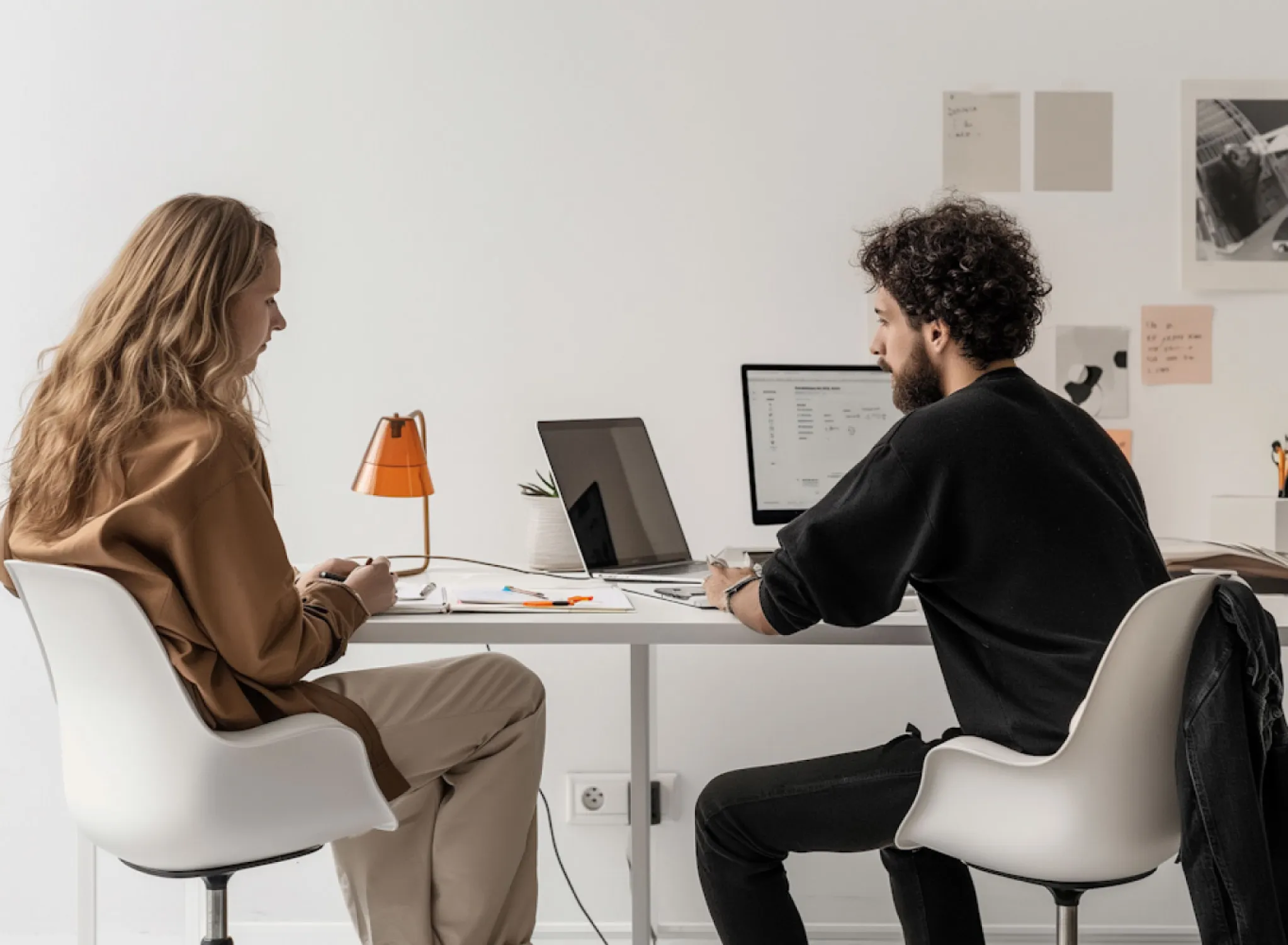 Two people sitting across from each other at a white table with laptops and papers, engaged in discussion in a modern office.