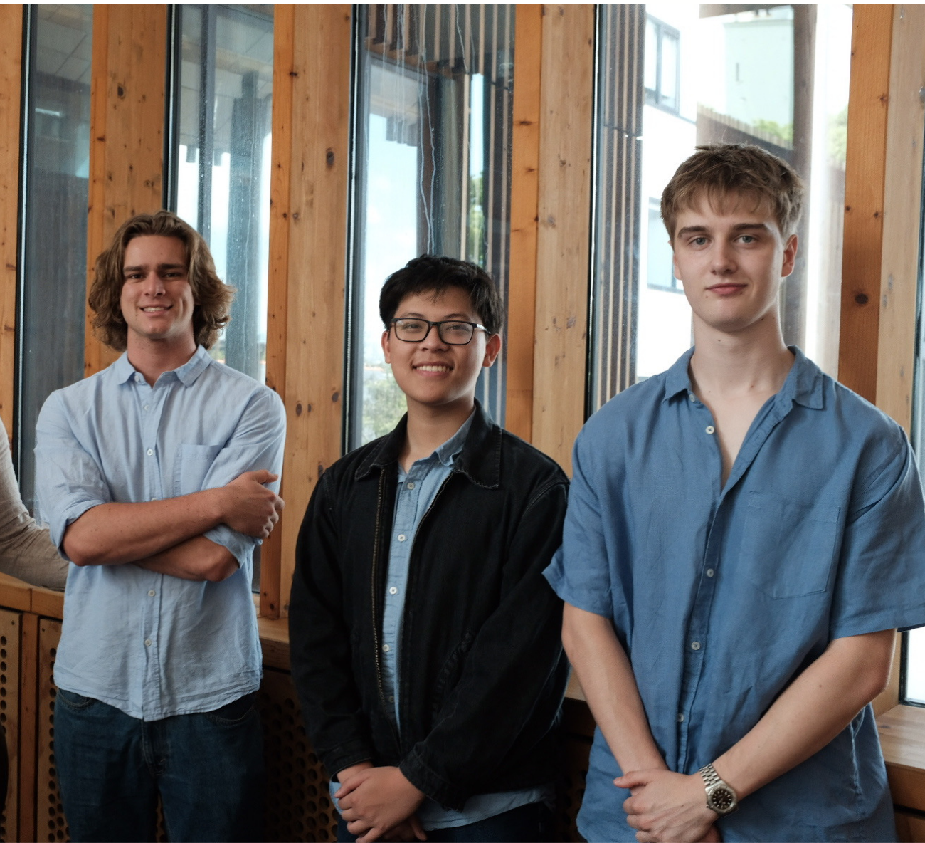 Three young men standing indoors in front of large wooden framed windows, smiling at the camera.
