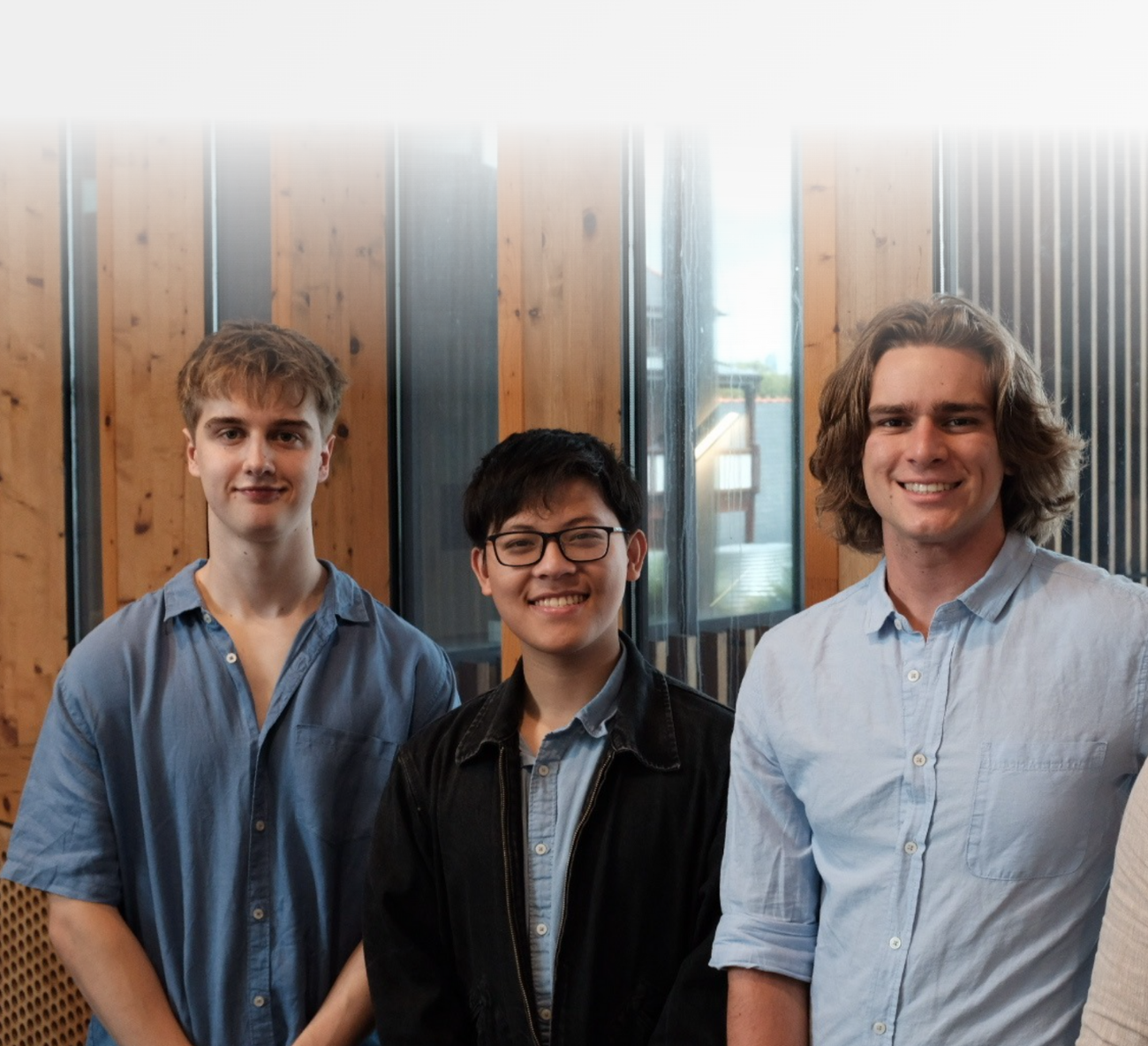 Three young men smiling and standing indoors in front of wooden panel walls and large windows.