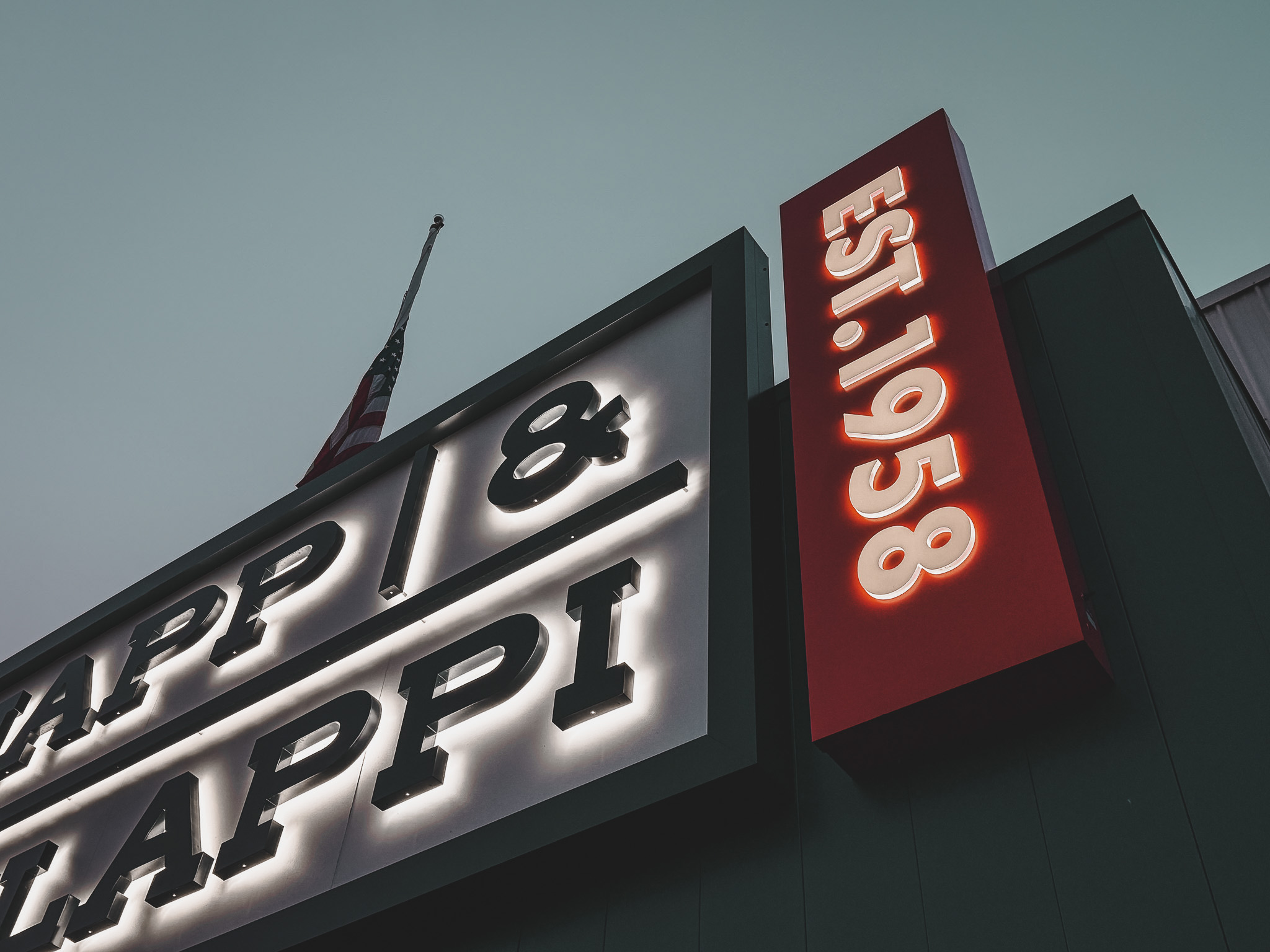 Illuminated outdoor sign for Zapp & Zappi with establishment year 1958 and an American flag at half-mast.