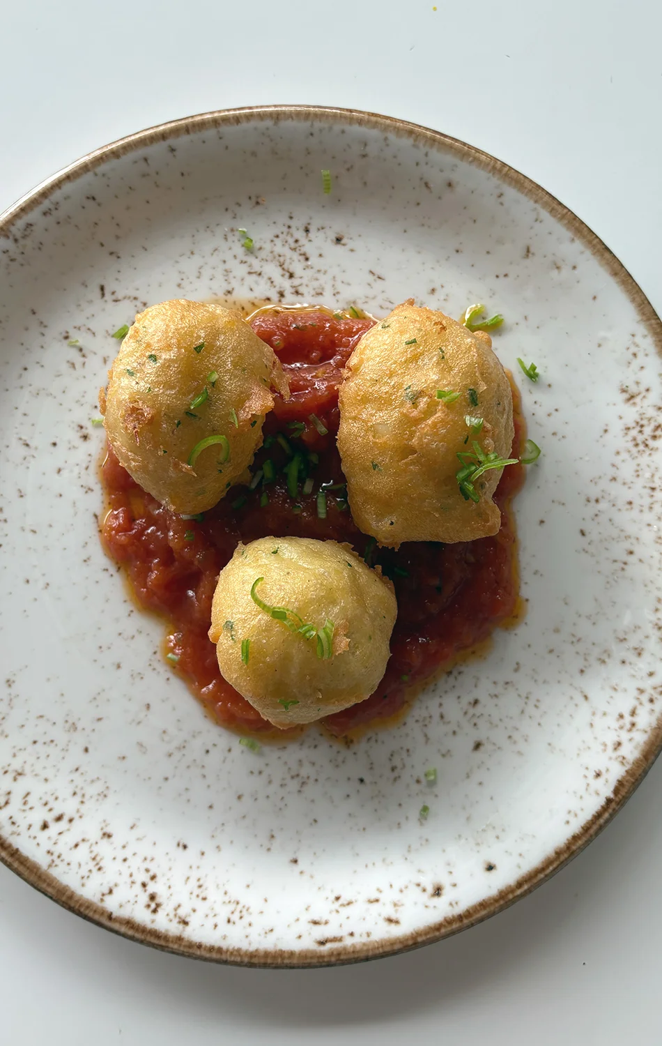 Three golden fried dough balls garnished with chopped herbs on a bed of red tomato sauce, served on a speckled white plate.