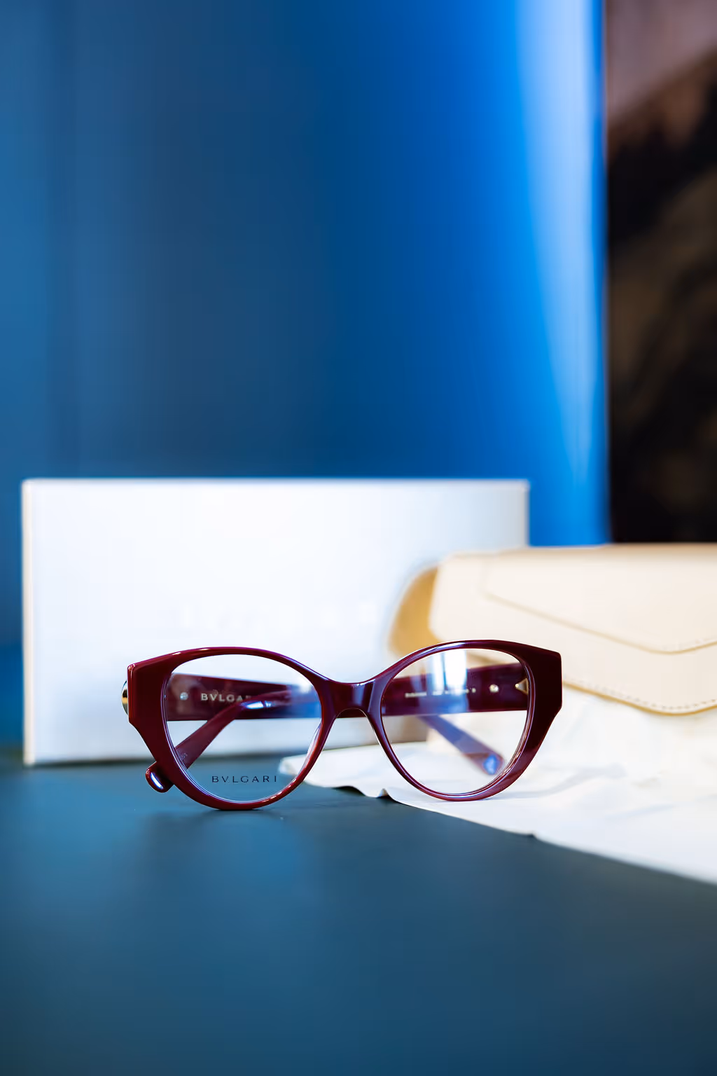 Burgundy Bvlgari cat-eye glasses on white surface with blue background