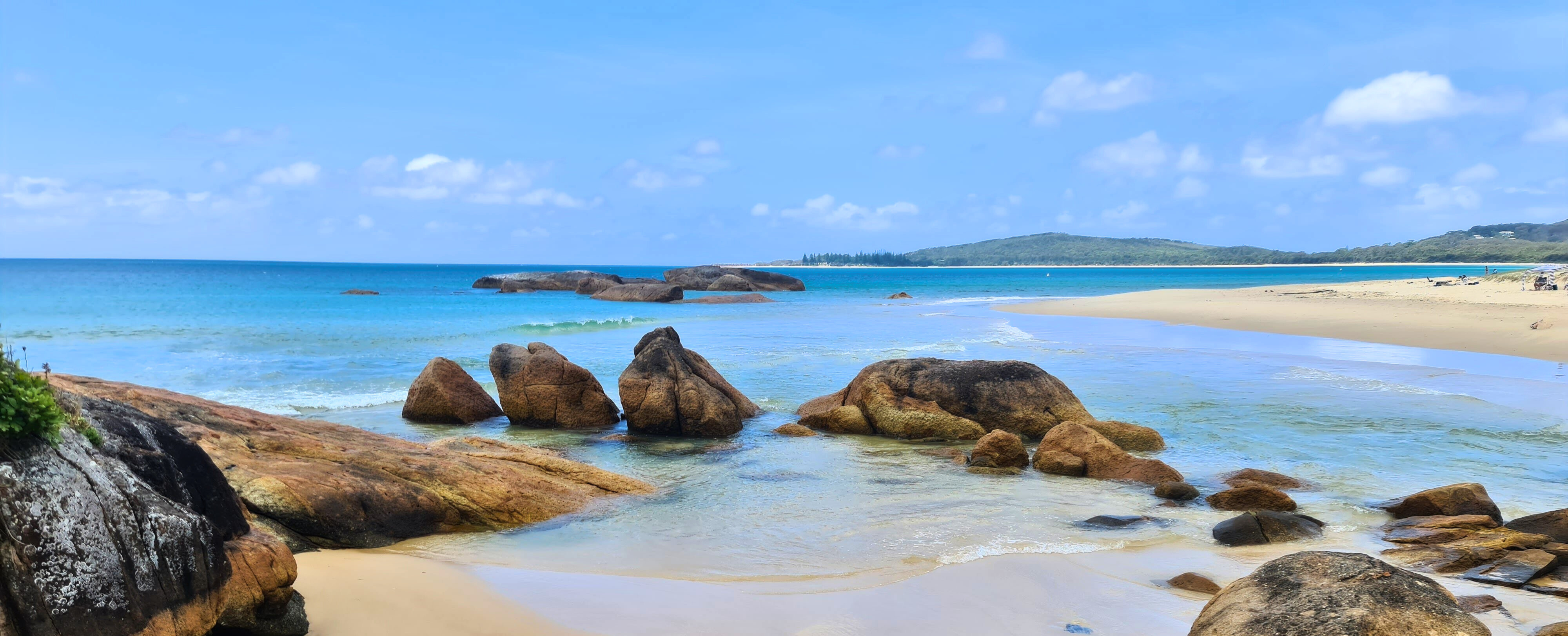 Calm turquoise ocean of Main Beach at South West Rocks, NSW, with boulders along a clean, sandy beach under a bright blue sky with scattered clouds.