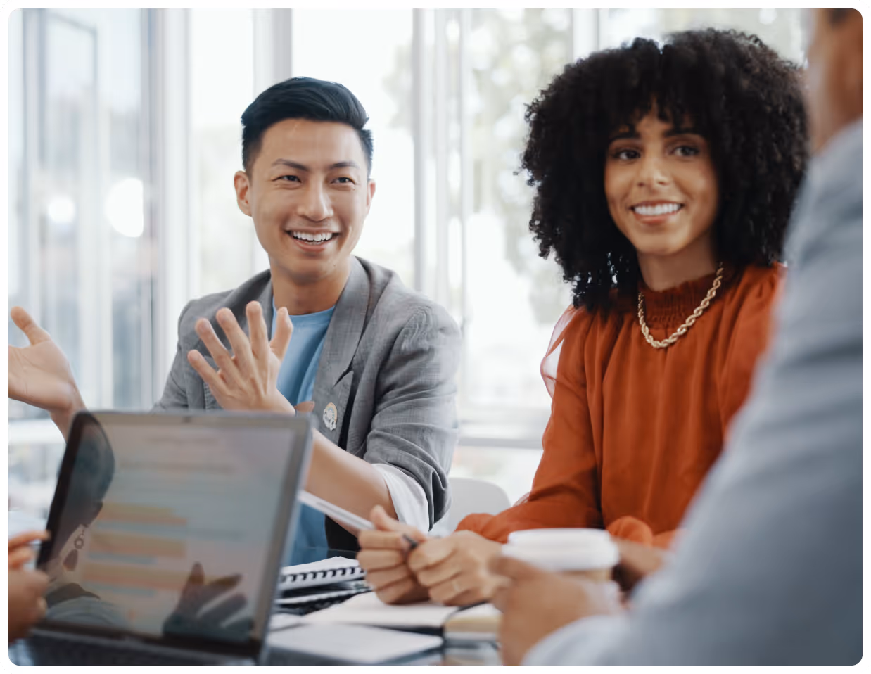 Two colleagues smiling and discussing work around a table with a laptop and notebooks.