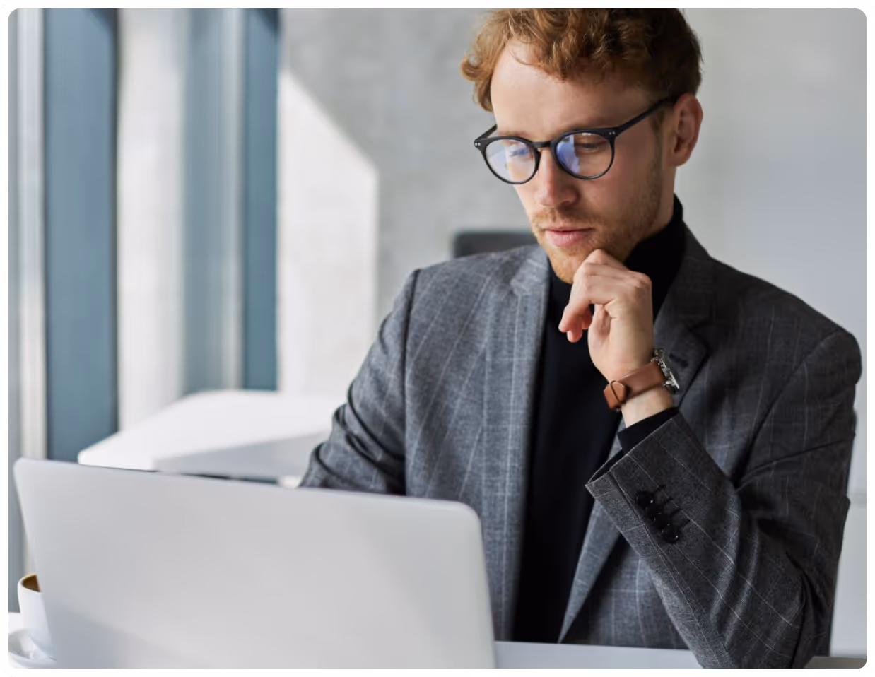 Man wearing glasses and a checked blazer working thoughtfully on a laptop indoors.