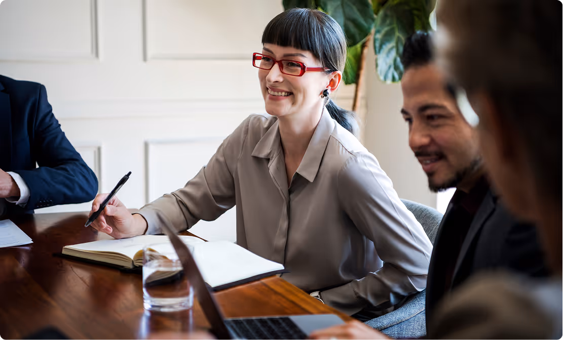 Smiling woman in red glasses writing in a notebook during a meeting with colleagues around a wooden table.