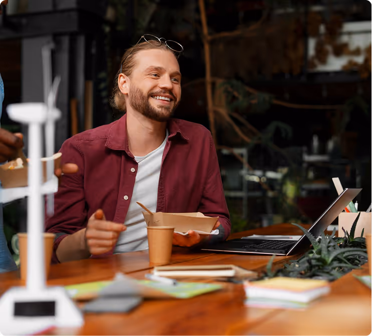Smiling man in a maroon shirt eating from a paper food container while sitting at a wooden table with a laptop and stationery.