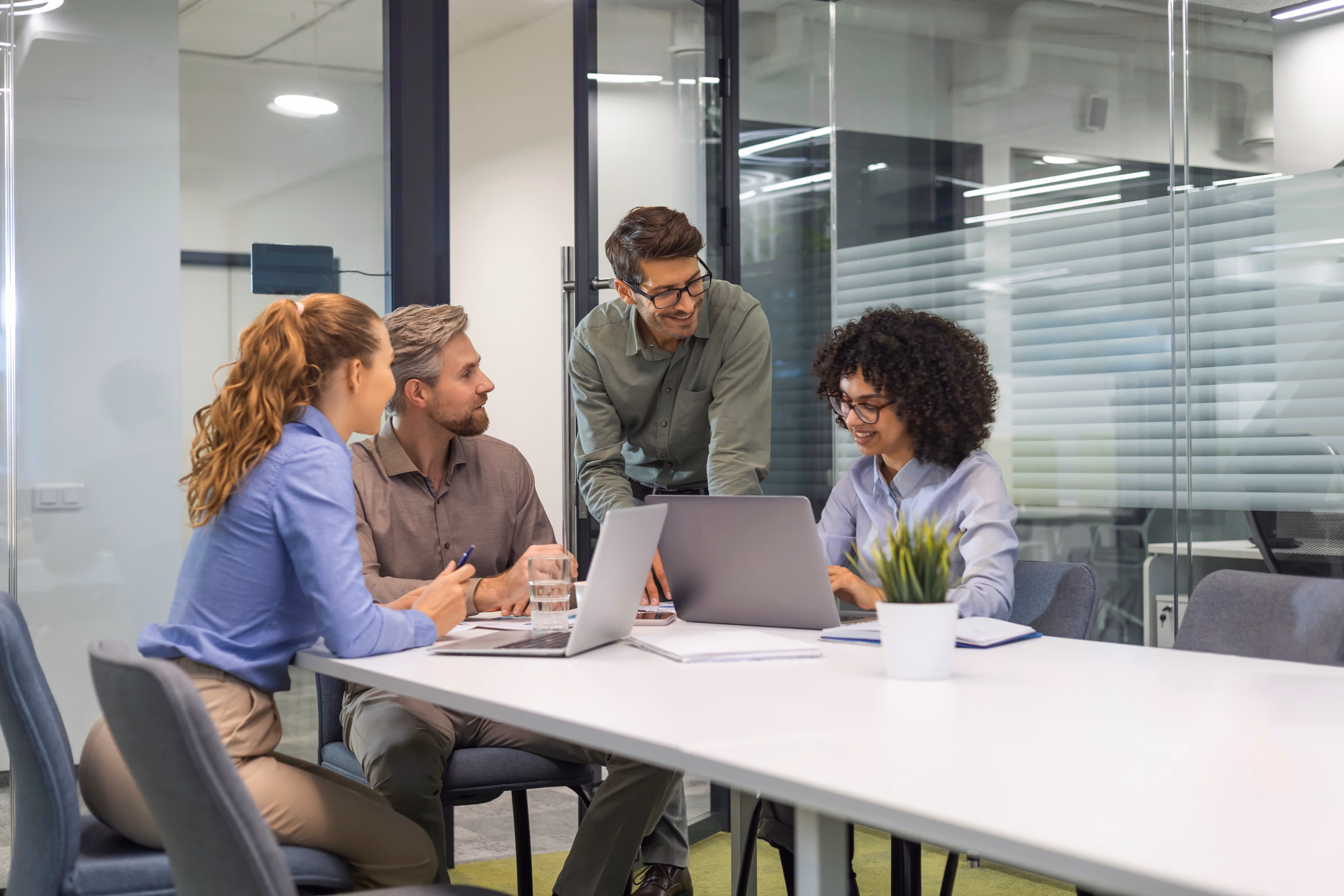 Four diverse colleagues collaborating at a table with laptops and notebooks in a modern office.