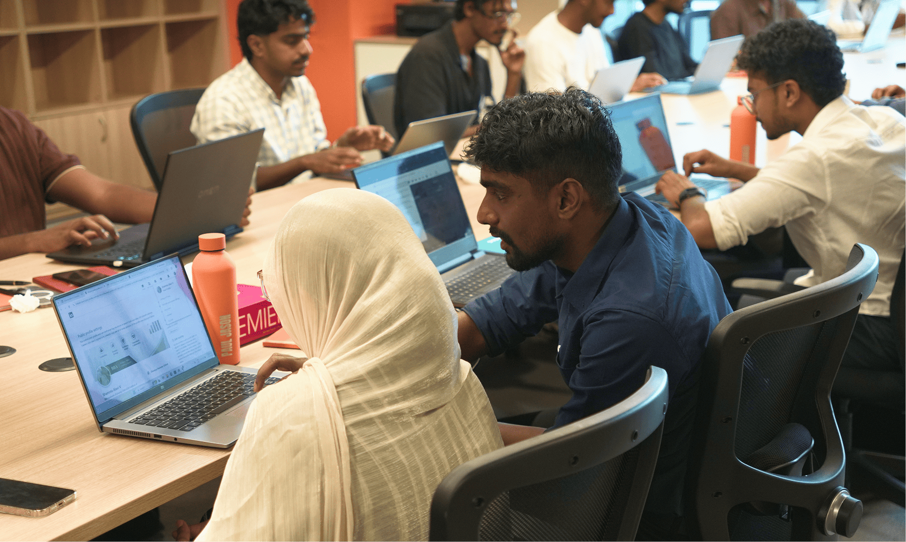 Students working side by side on laptops at a shared table, learning through hands-on projects rather than a traditional classroom lecture.