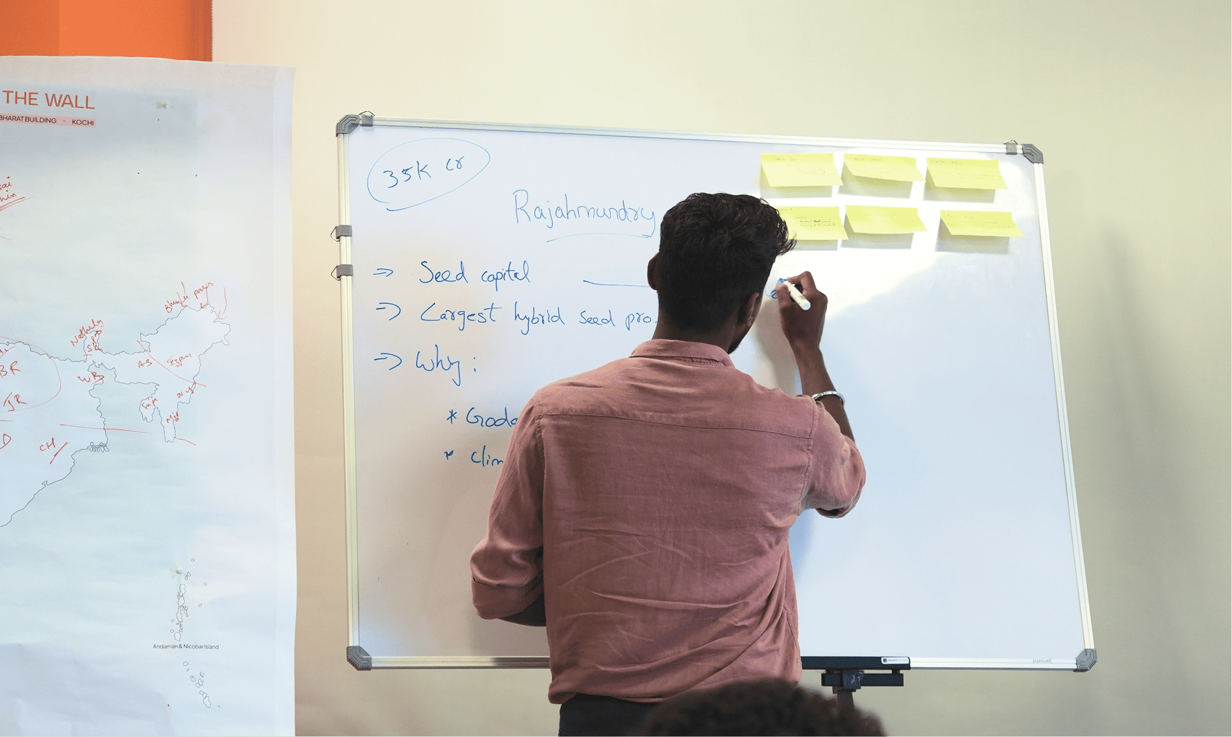 A student writing a business case on a whiteboard, with sticky notes above and an annotated map of India on the wall behind.