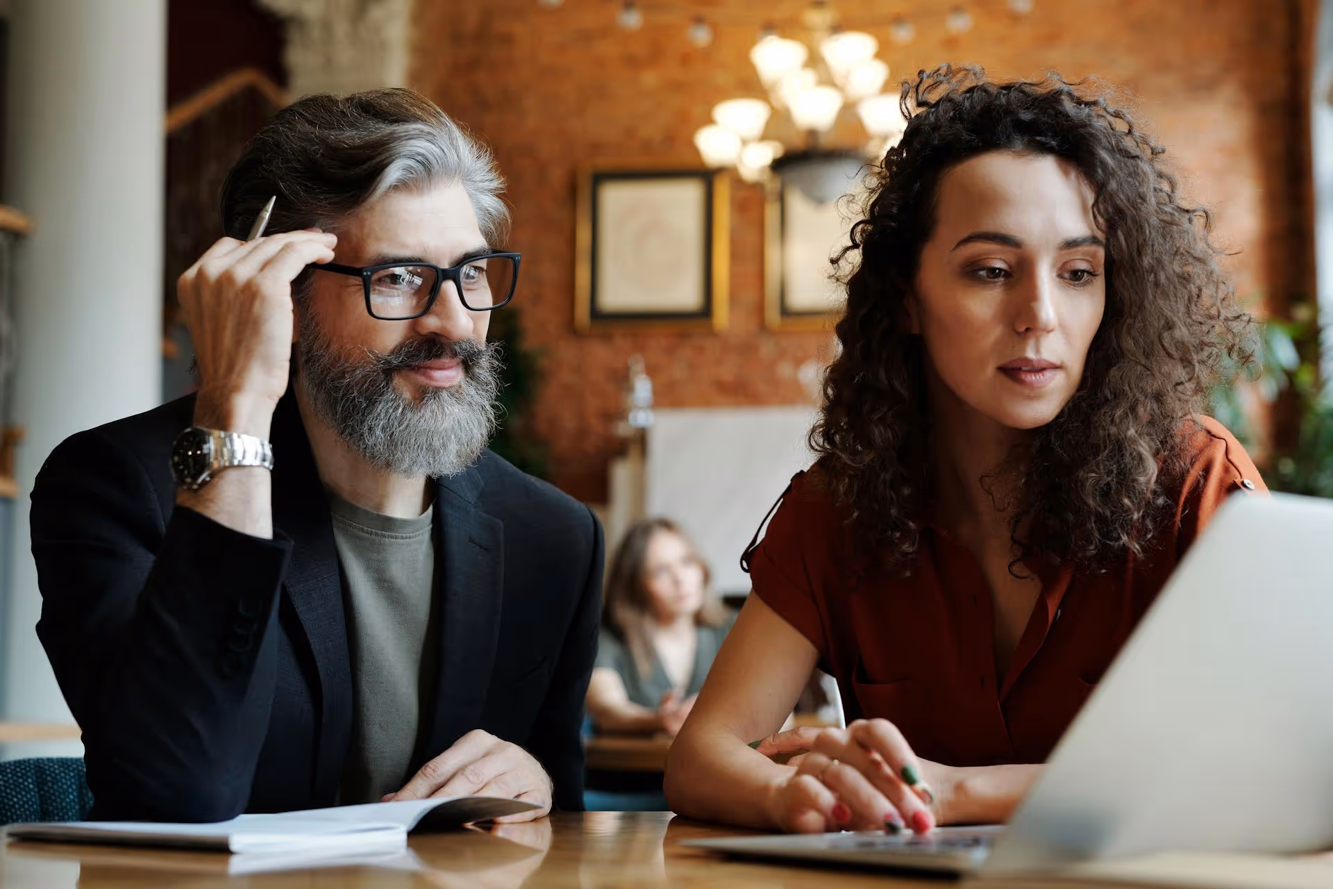 Man with gray beard and glasses and woman with curly hair working together on a laptop in a café.