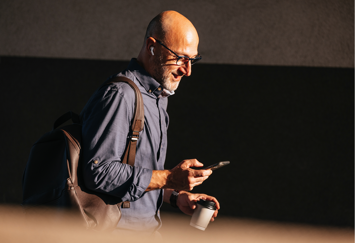 Bald man with glasses walking, holding coffee cup and looking at his phone with earbuds in ears.