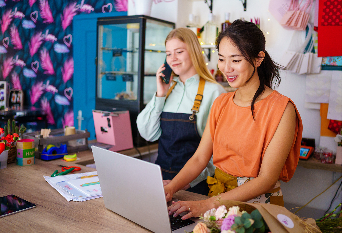 Two women working in a flower shop with laptop and phone, surrounded by floral and crafting supplies.