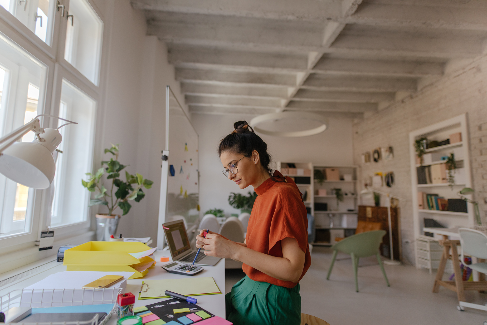 Woman in glasses and orange shirt multitasking with makeup and laptop in a bright office.