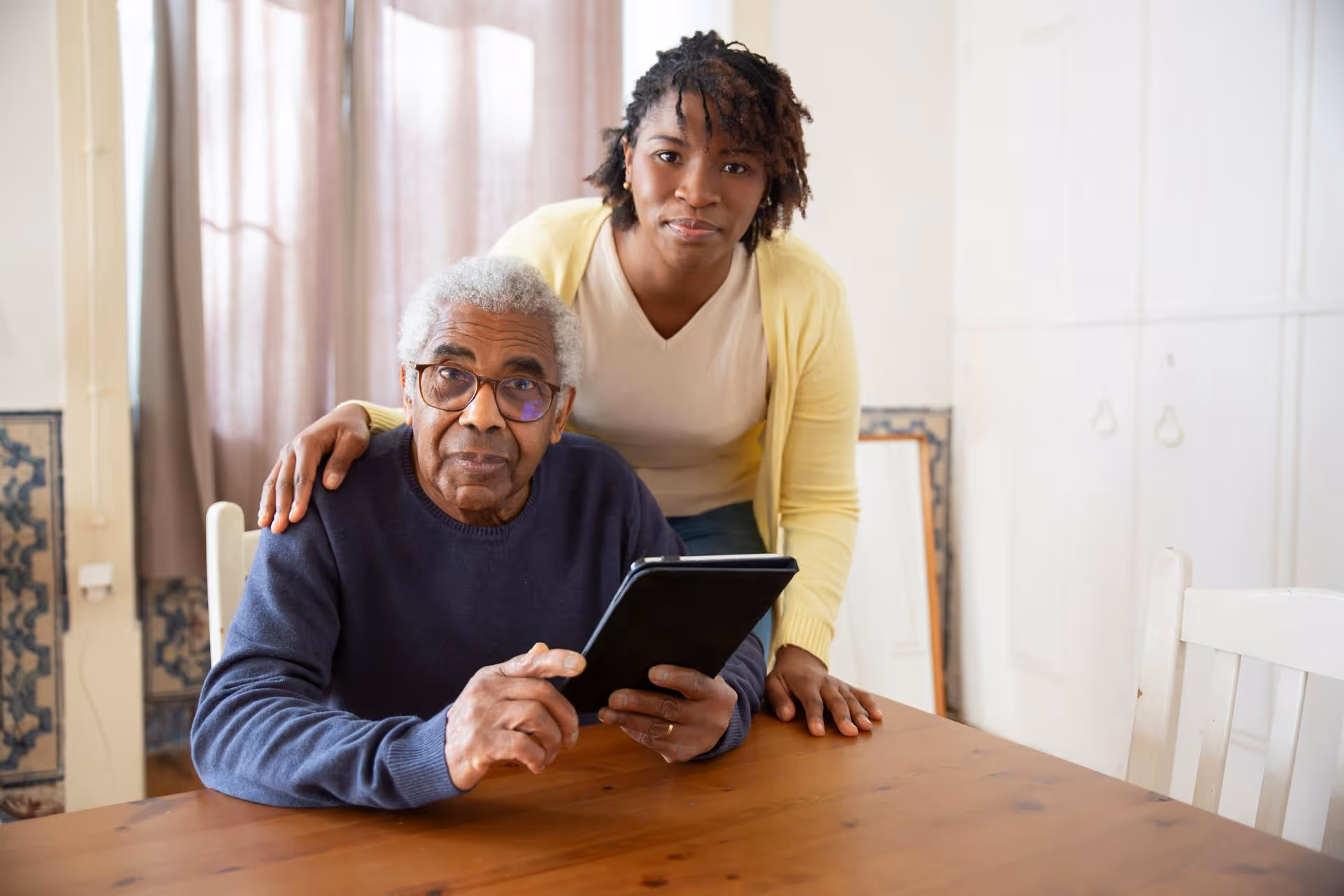 Elderly man with glasses holding a tablet, younger woman standing behind him with a hand on his shoulder.