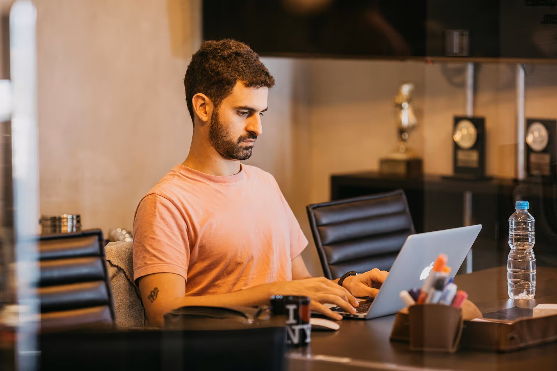 Man in pink shirt working on a laptop at a conference table with a water bottle and pens nearby.