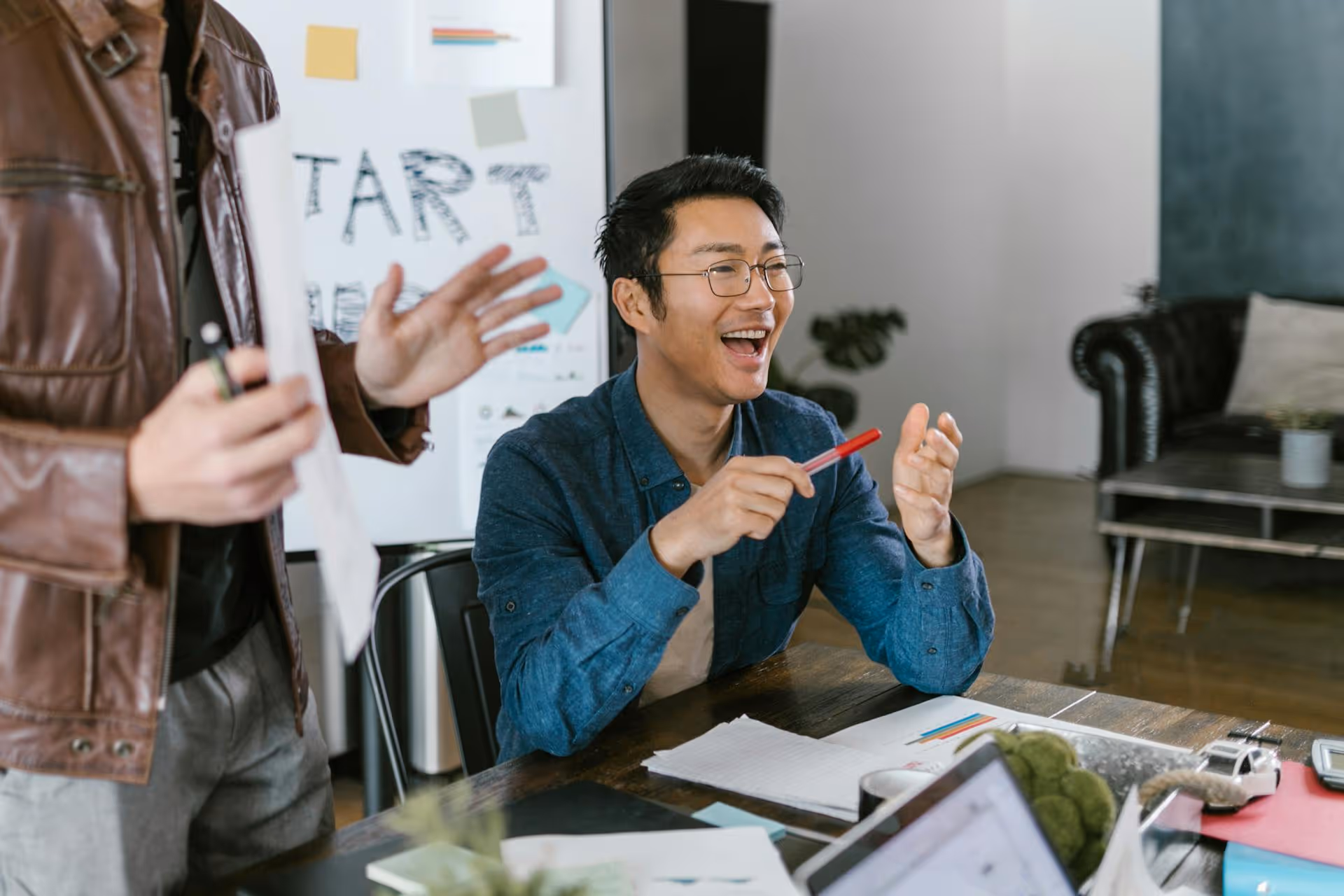 Man in glasses laughing and holding a pen sitting at a table during a meeting or brainstorming session.