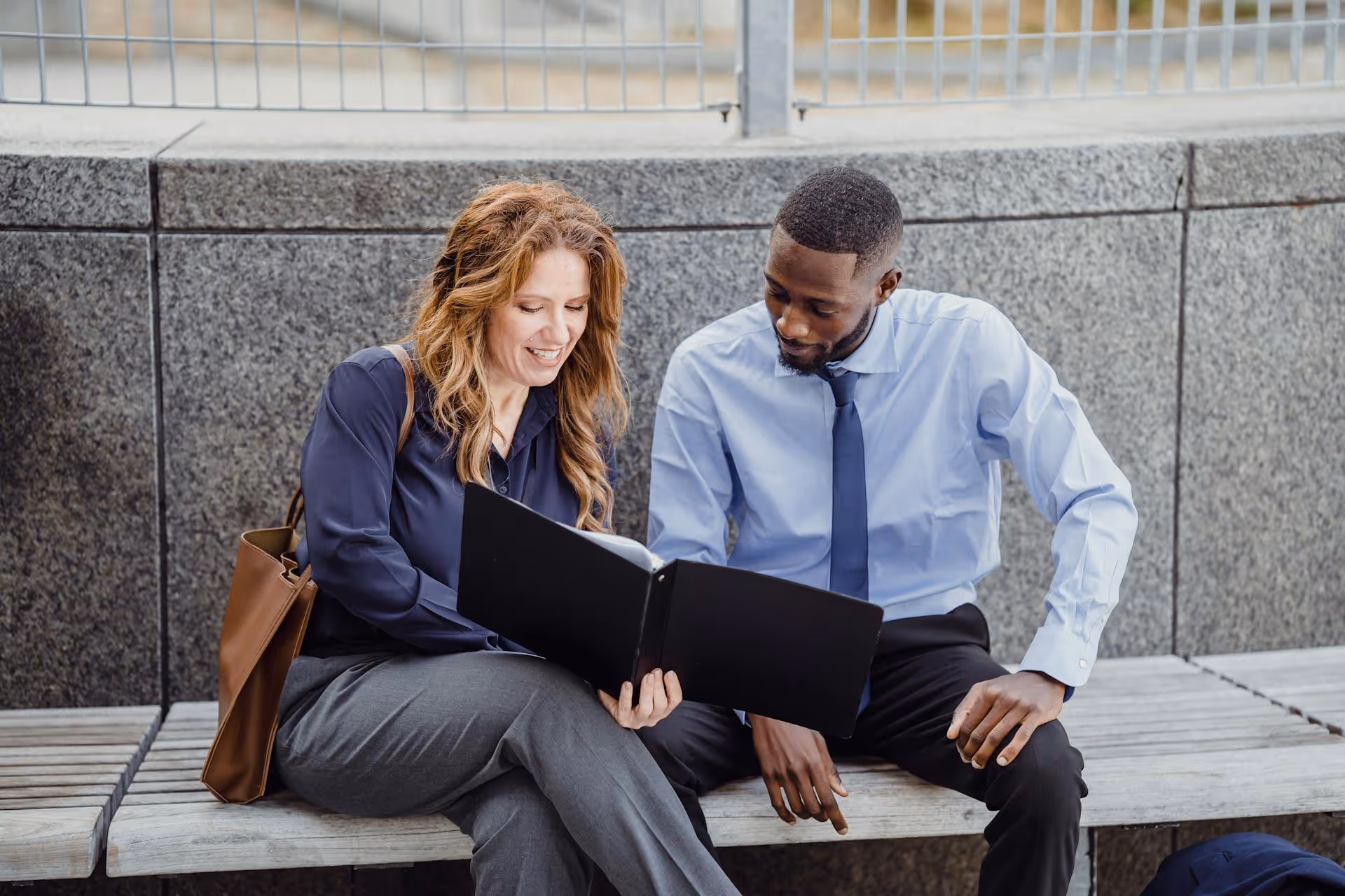 Two business professionals sitting on a bench reviewing documents in a black folder together.
