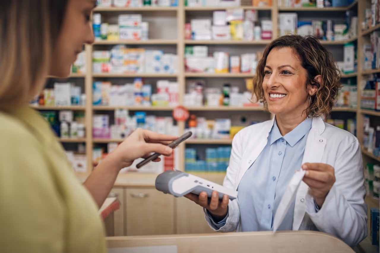 Pharmacist in white coat smiling while holding receipt and card reader as customer pays by phone.