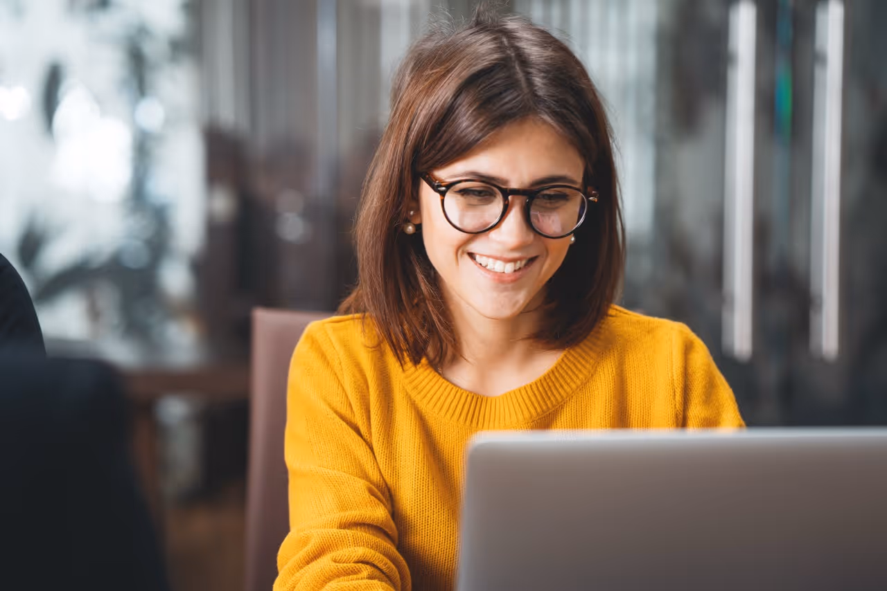 Smiling woman with glasses in yellow sweater looking at a laptop in an office setting.