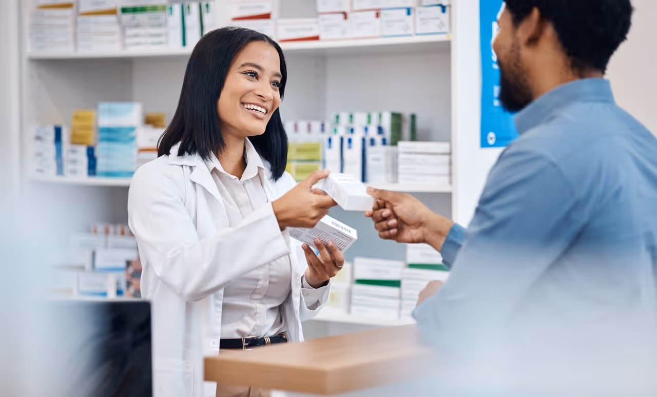 Pharmacist smiling while handing medication boxes to a customer at a pharmacy counter.
