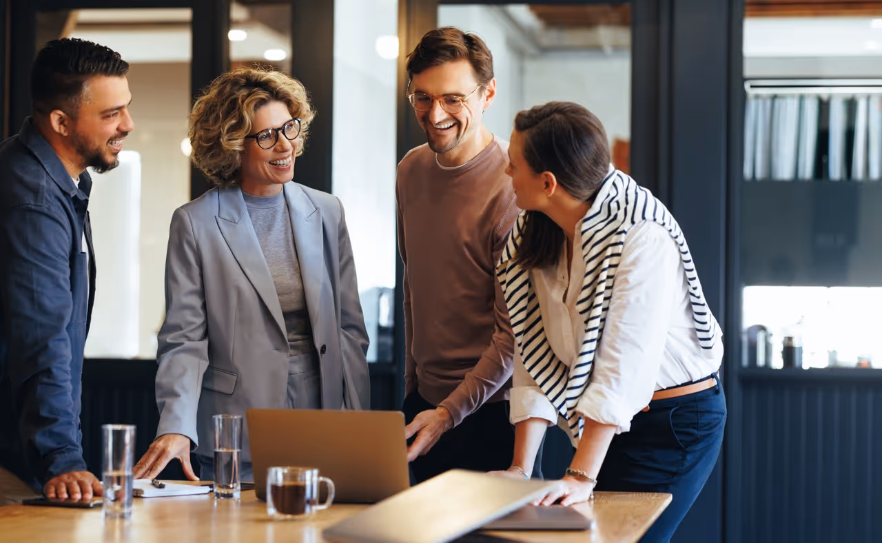 Four coworkers smiling and discussing around a desk with a laptop in a modern office.