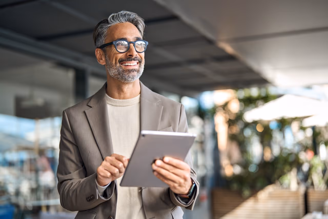 Smiling middle-aged man with gray hair and glasses using a tablet in a modern indoor setting.