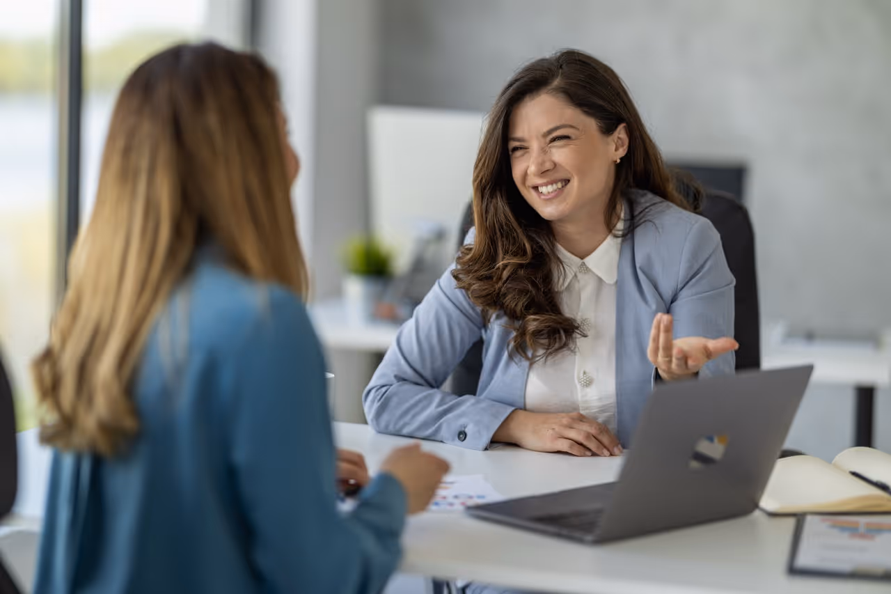Woman in blue blazer smiling while talking to another woman at a desk with a laptop.