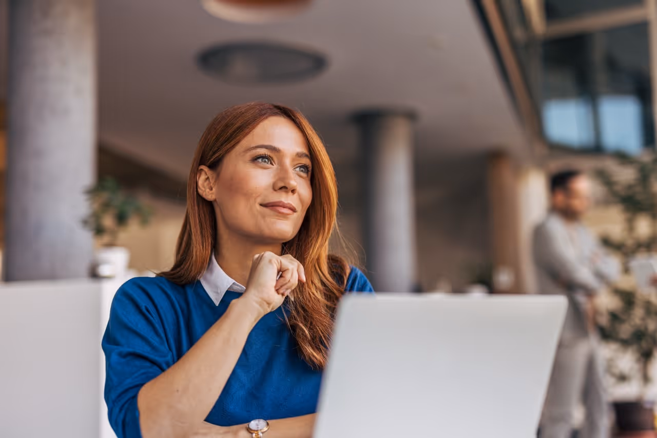 Woman in blue sweater sitting at a laptop, looking thoughtfully to the side indoors.