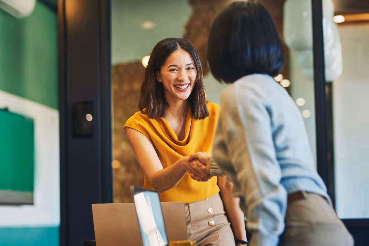 Two women smiling and shaking hands in a modern office setting.