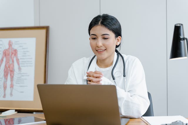Female doctor wearing a stethoscope, sitting at a desk, smiling while looking at a laptop.