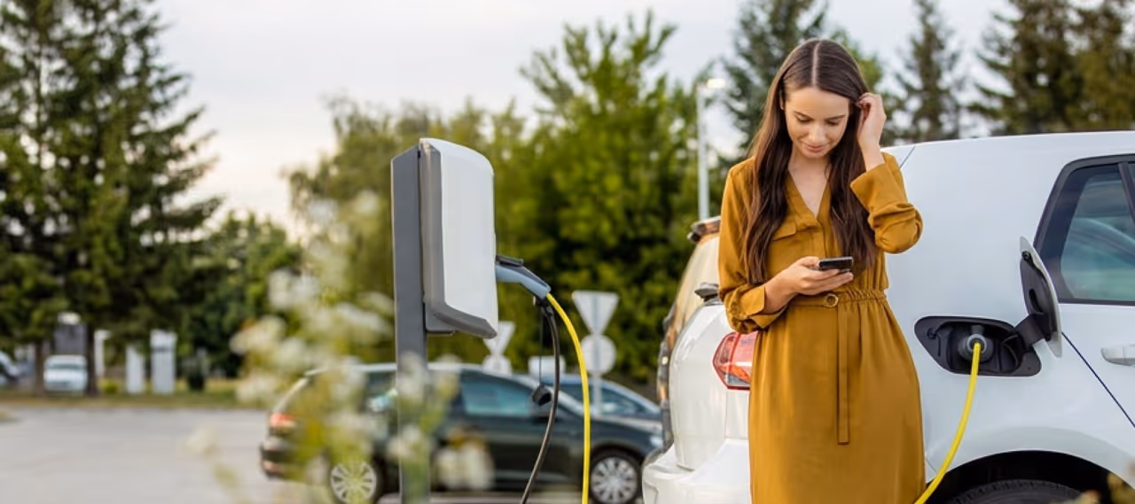 Young woman in a brown dress using her phone while charging a white electric car outside.