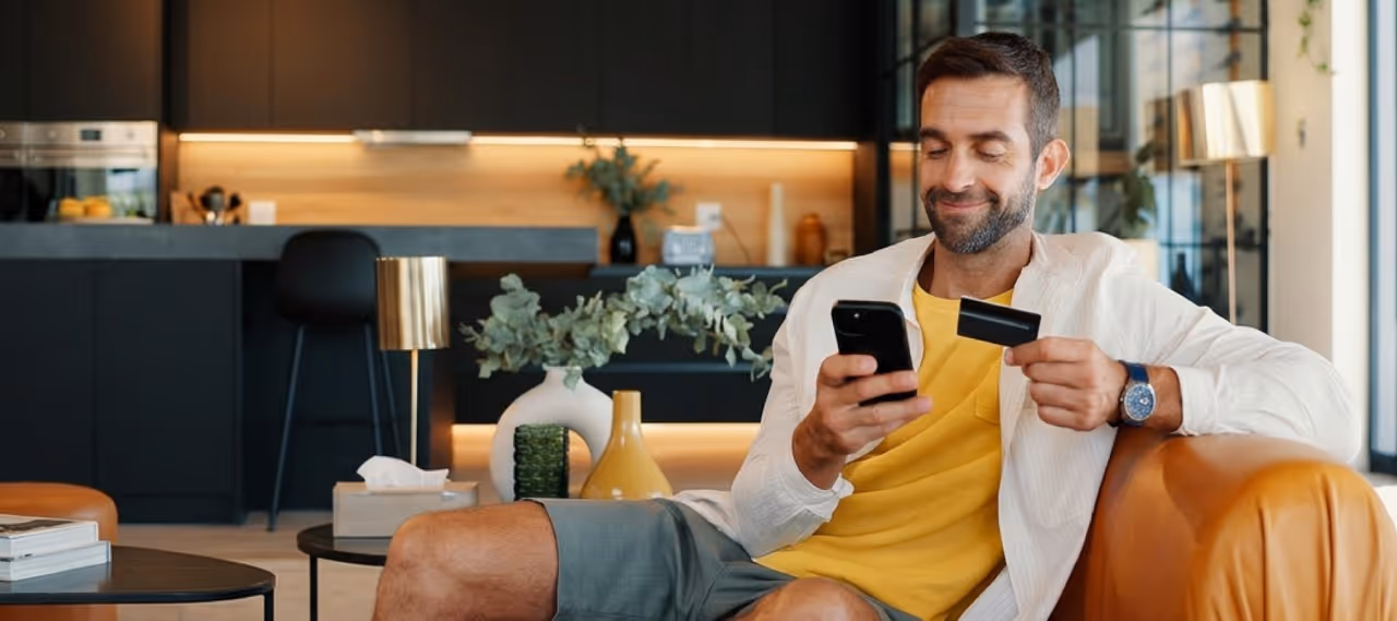 Smiling man in yellow shirt and white jacket holding credit card and phone while sitting on sofa.