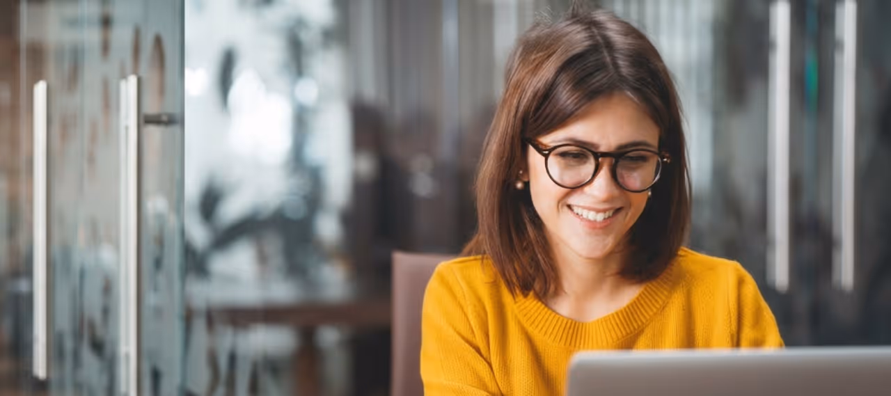 Smiling woman wearing glasses and a yellow sweater working on a laptop indoors.