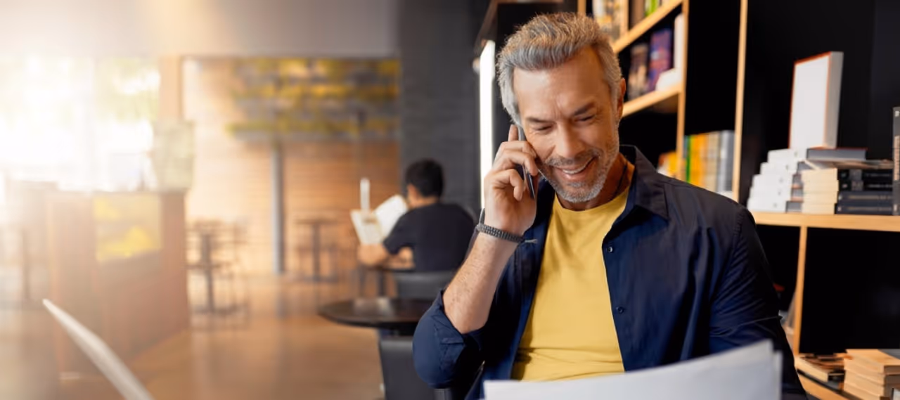 Smiling man in a bookstore talking on phone while reading a newspaper or document.