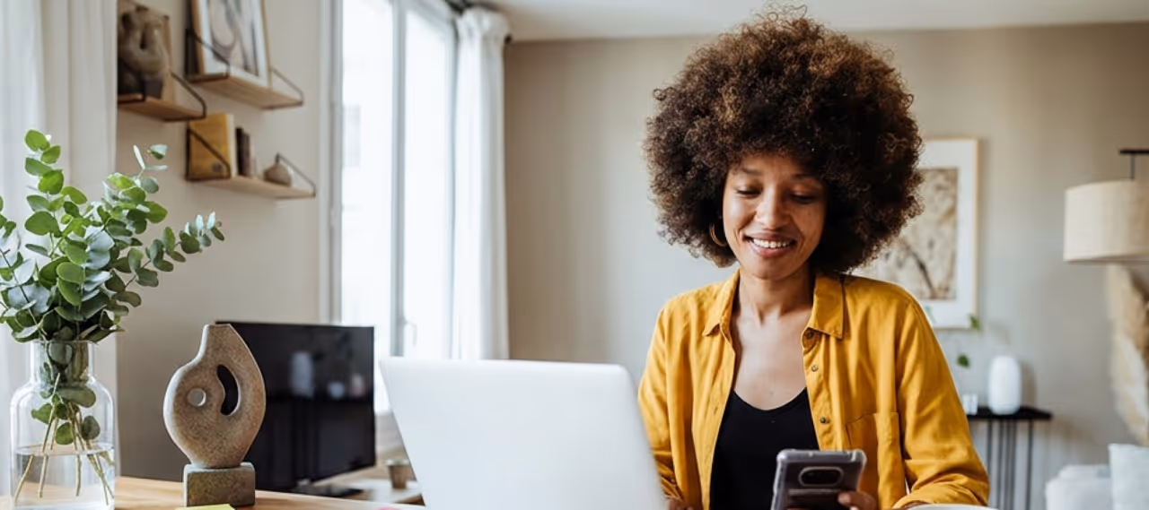 Woman with curly hair in a yellow shirt smiling while using phone and laptop at home desk.
